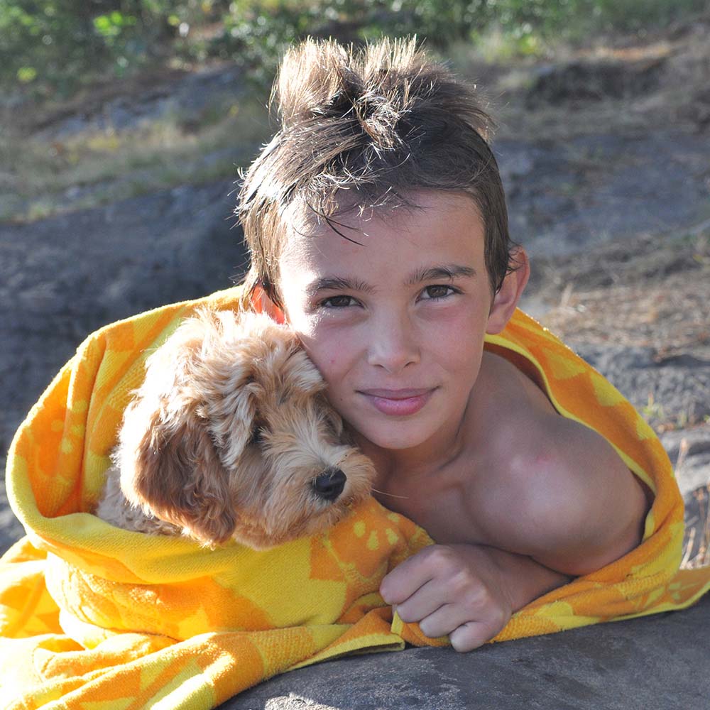 Smiling boy cuddling his fluffy dog wrapped in a bright yellow towel after swimming, showing warmth and companionship outdoors.
