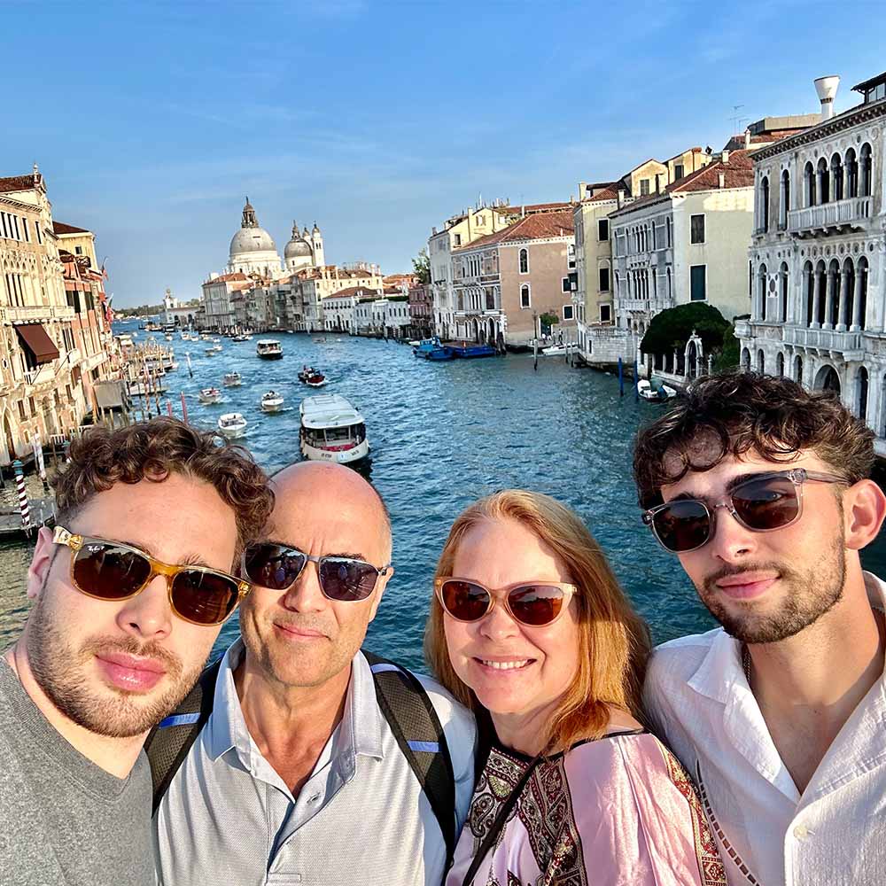 Glowhound founders smiling on a Venice canal bridge with scenic waterway, historic buildings, and boats in the background.