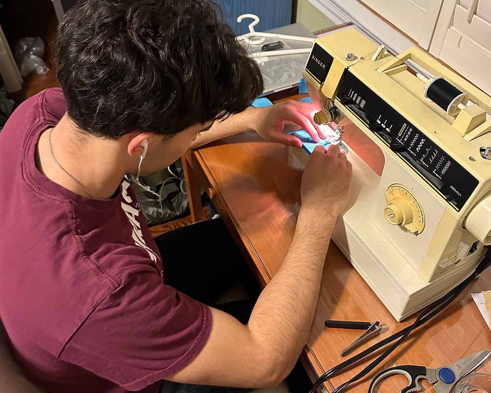 Young man listens to music while sewing blue fabric on a vintage Singer sewing machine at a wooden desk in a cozy room.